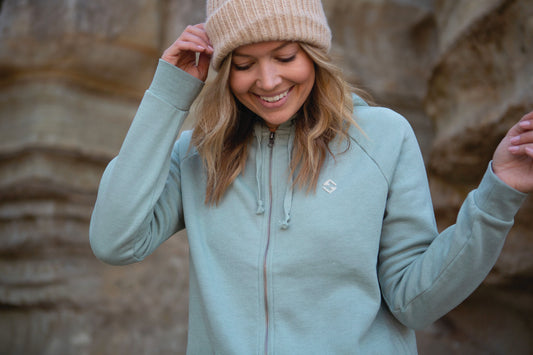 Woman wearing a light green hoodie and beige beanie standing against a rocky background
