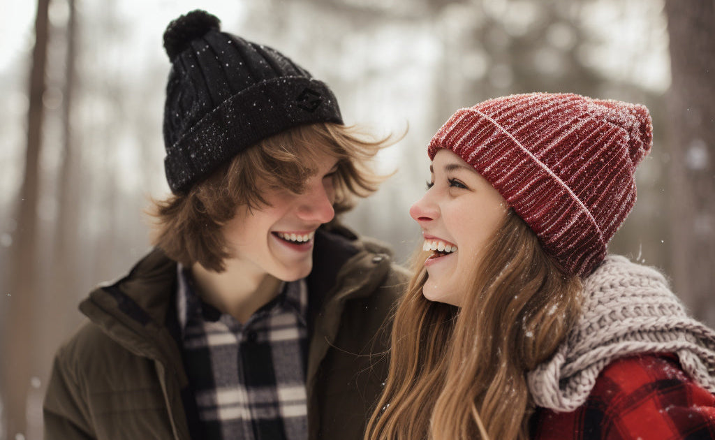 Two people in winter clothing smiling at each other in a snowy forest.