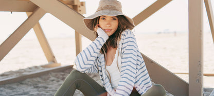 Zip-Up Hoodie – Heathered French Terry Woman sitting on sand wearing a striped shirt and green leggings with a straw hat.
