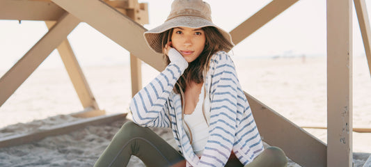Woman sitting on sand wearing a striped shirt and green leggings with a straw hat.
