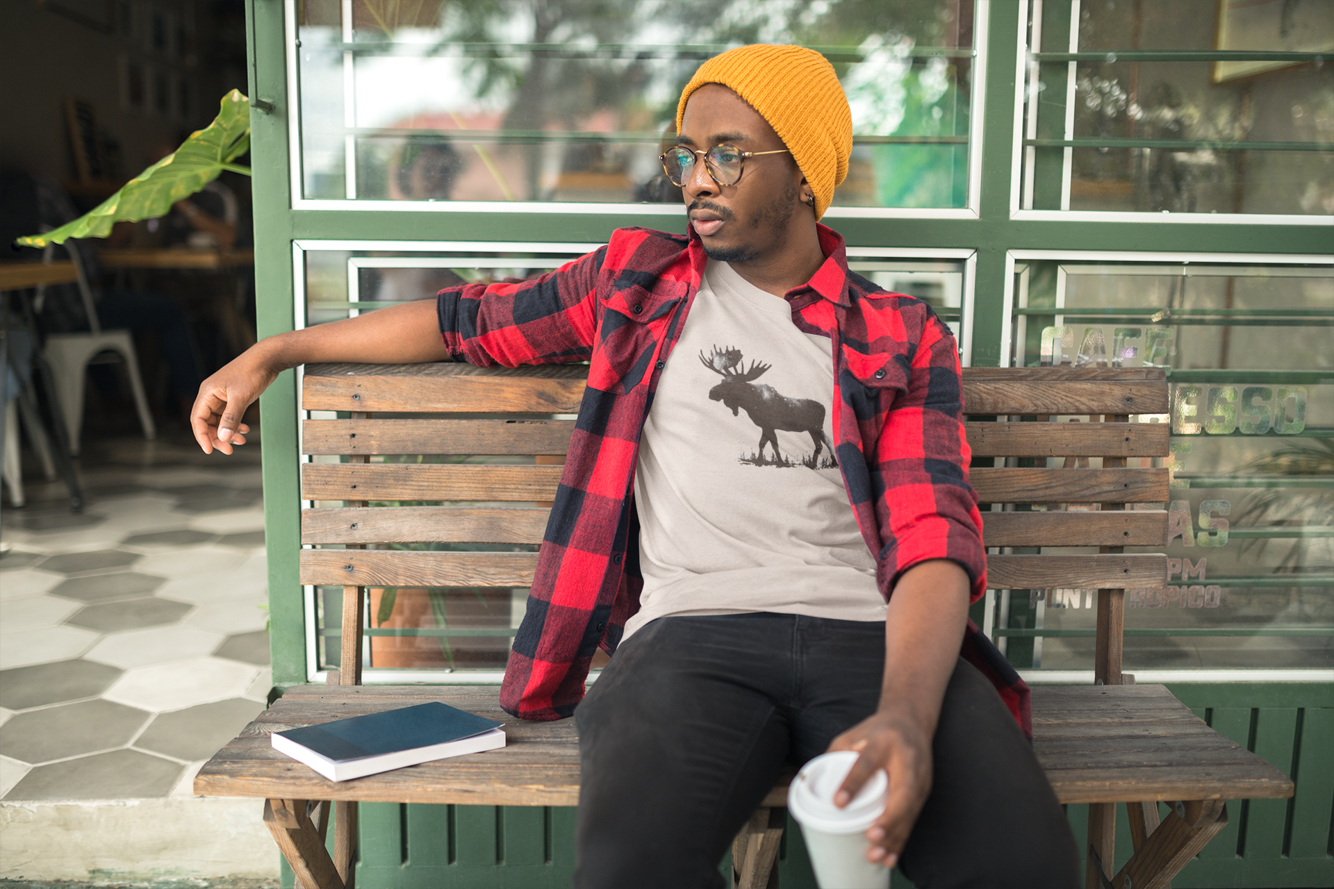 Person sitting on a bench wearing a red plaid shirt and yellow beanie, with a moose graphic on a white t-shirt.